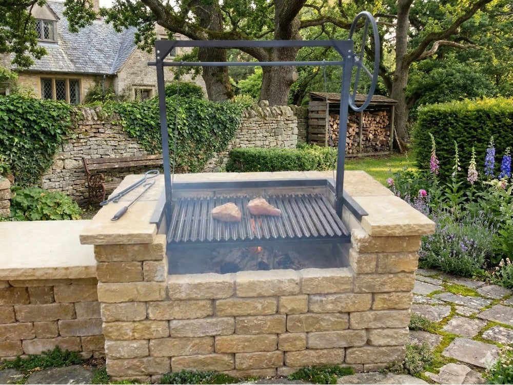 Argentine grill kit with 3-sided flange and adjustable wheel height system installed in a masonry firebox, grilling steaks over a wood fire in an outdoor kitchen setting.