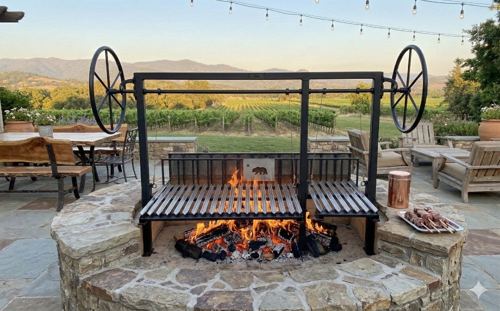 Split Argentine grill kit with dual adjustable grates and rear braseros installed in a stone outdoor kitchen fire pit with wood-fire cooking.