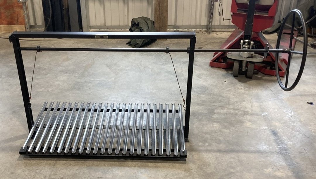 Black steel Argentine bolt-in grill kit with stainless V-grate and extended wheel control rod designed to pass through a masonry wall for use in an open-front fireplace installation.