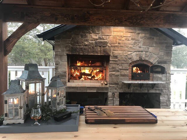 Argentine-style grill with rear brasero installed in a stone outdoor kitchen, featuring a live fire cooking area with burning wood inside a covered backyard patio