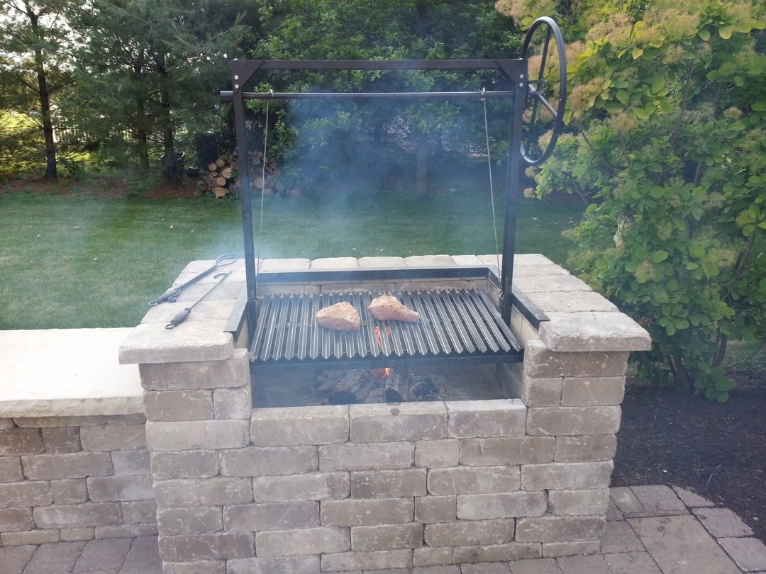 Model #4546 Argentine architectural grill with 3-sided flange installed in a stone masonry firebox, cooking steaks over a wood fire with adjustable wheel height control.