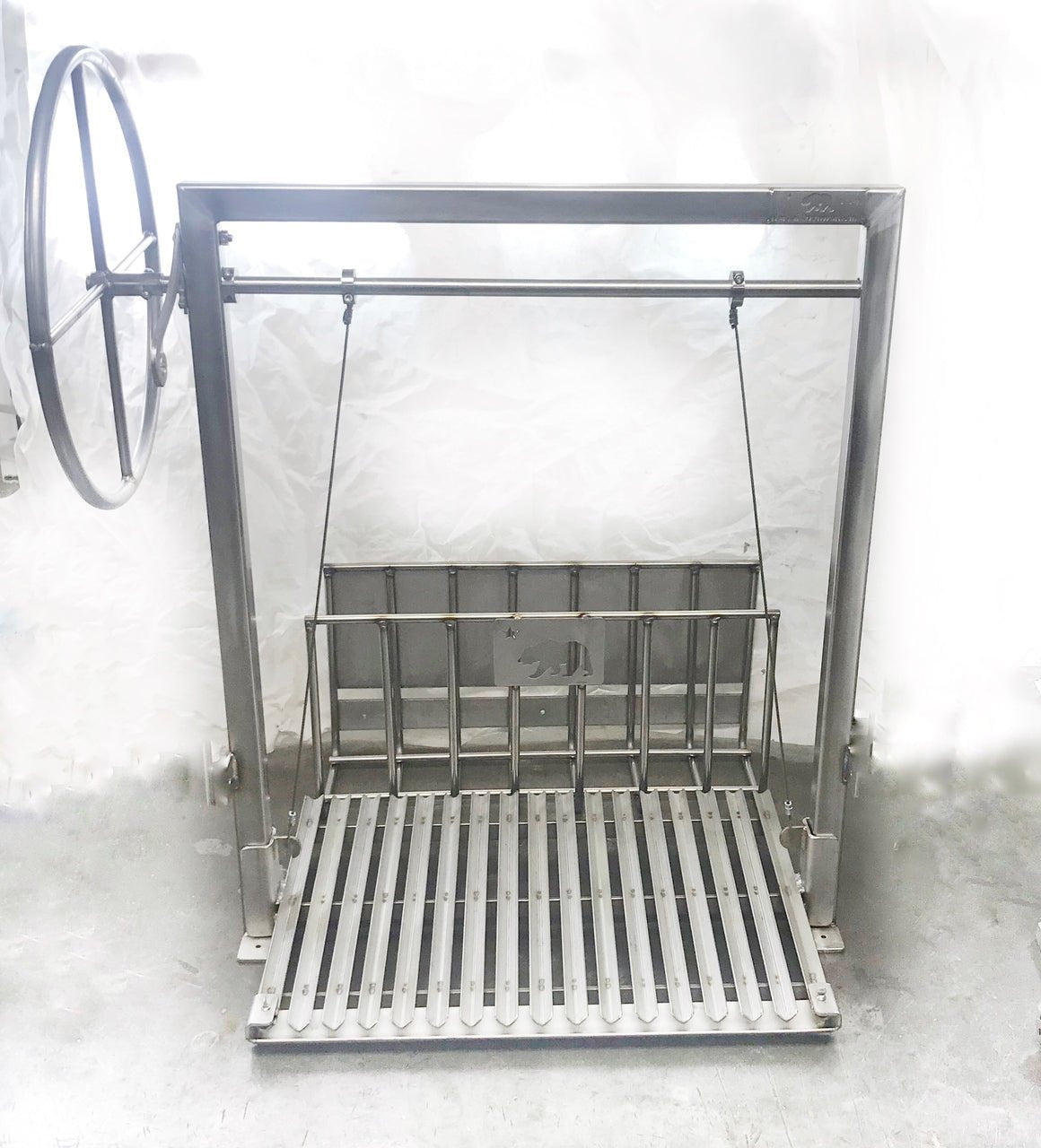 Argentine-style stainless steel grill with rear brasero and adjustable height grate, featuring a side wheel control and round rod cooking surface for wood or charcoal grilling