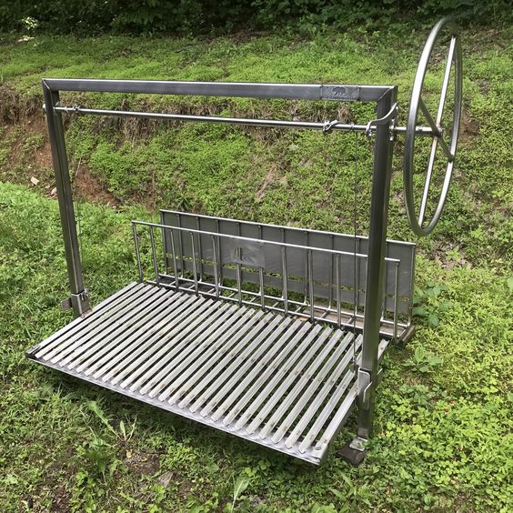 Argentine-style grill with rear brasero and adjustable-height grate, featuring a side wheel control and a round rod cooking surface, shown outdoors on grass.