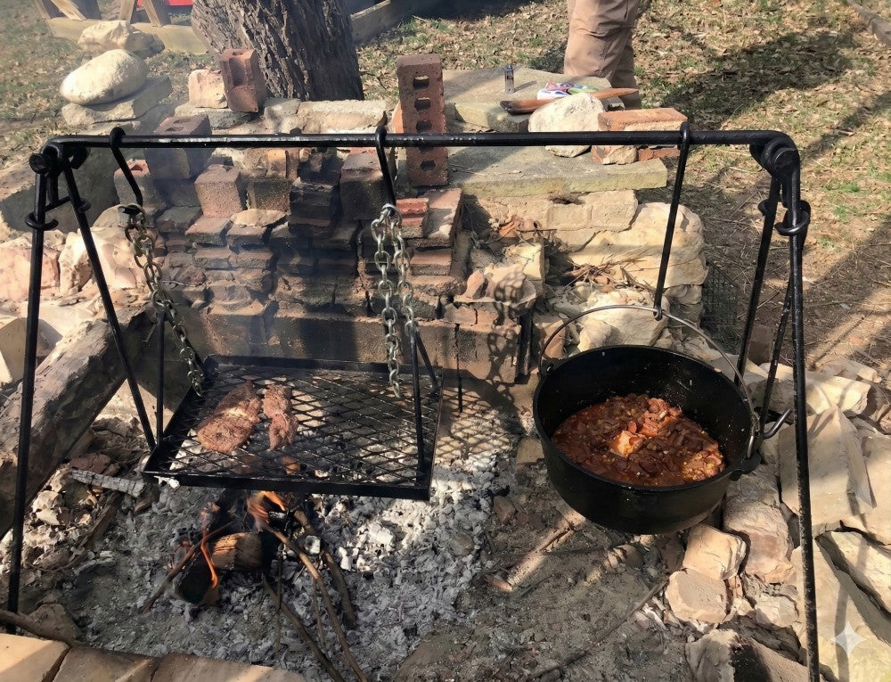 Dutch oven cooking chili beside grilled meat over a campfire using a quadpod outdoor cooking system.