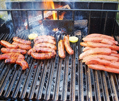 Sausages grilling over hot coals on an Argentine V-grate with a rear brasero burning wood in the background for authentic wood-fired cooking.
