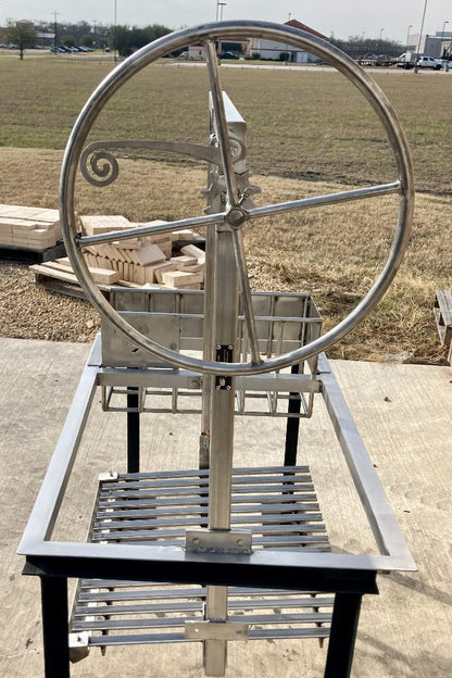 Side angle of stainless Argentine grill kit featuring the slanted V-grate, brasero firebox, and decorative hand wheel lift mechanism.