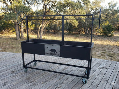 Split Santa Maria Style Grill by Heritage Backyard. Freestanding charcoal and wood grill with independent dual lift mechanisms, side wheels, and decorative bear emblem.