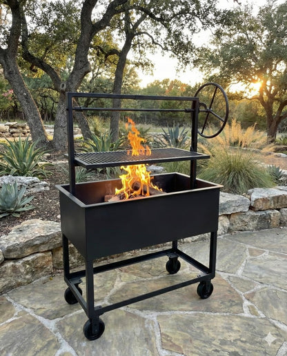 Portable Santa Maria–style grill with adjustable height grate and side wheel, featuring an open firebox with burning wood, set on a wheeled steel cart in an outdoor stone patio surrounded by oak trees and desert landscaping.