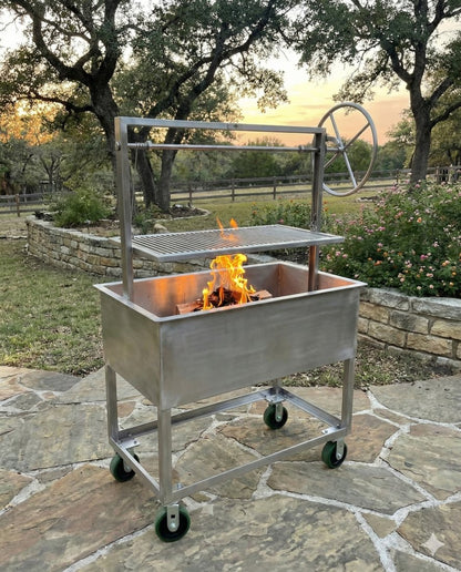 Portable stainless steel Santa Maria–style grill with adjustable height grate and side wheel, featuring an open firebox with burning wood, mounted on a wheeled cart and set on a stone patio with trees and garden landscaping at sunset.