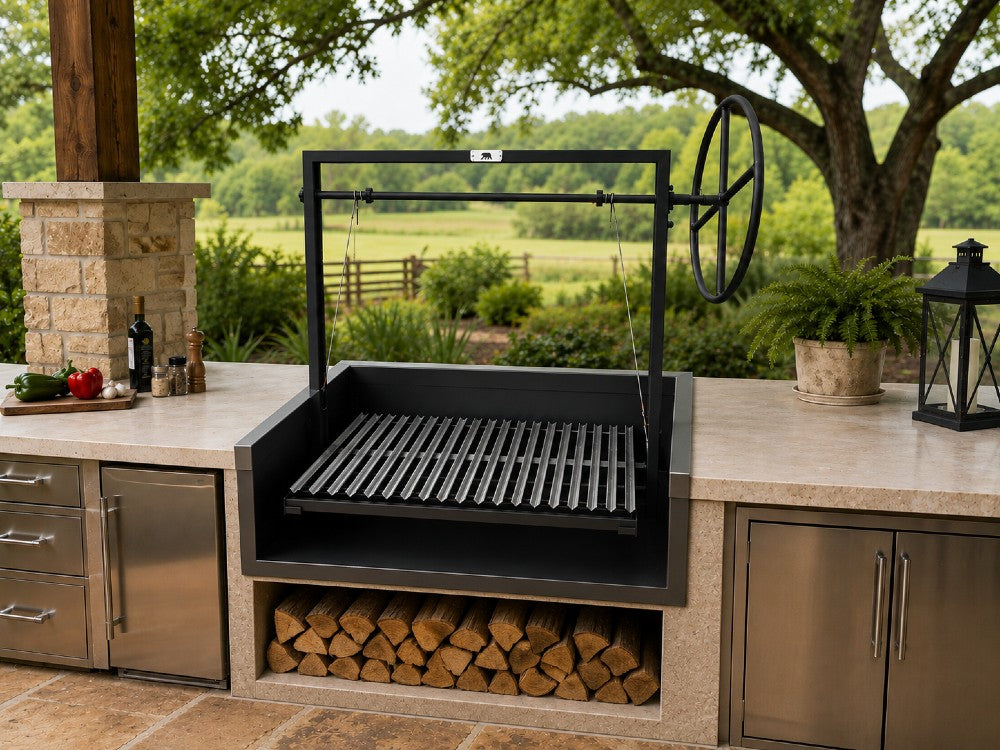 Open Front Argentine Built-In Grill with a black steel firebox and adjustable stainless steel V-grate installed in a custom outdoor kitchen with countertop flange mounting and integrated firewood storage below.