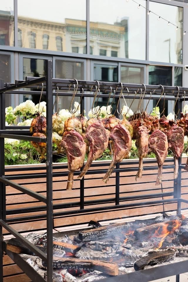 Tomahawk steaks roasting on hanging hooks over open flames on Portico BBQ grill